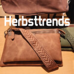 Braune und olivgrüne Umhängetaschen mit breiten, dekorativen Schulterriemen auf einer Verkaufsfläche, darüber der Schriftzug Herbsttrends.