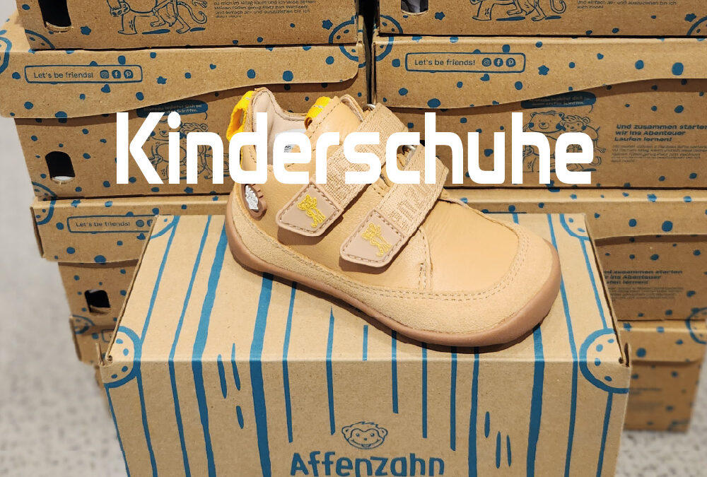 Beiger Affenzahn Kinderschuh mit zwei Klettverschlüssen und brauner Sohle auf einem Schuhkarton, vor gestapelten Kartons mit dem Schriftzug Kinderschuhe.