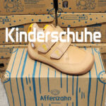 Beiger Affenzahn Kinderschuh mit zwei Klettverschlüssen und brauner Sohle auf einem Schuhkarton, vor gestapelten Kartons mit dem Schriftzug Kinderschuhe.