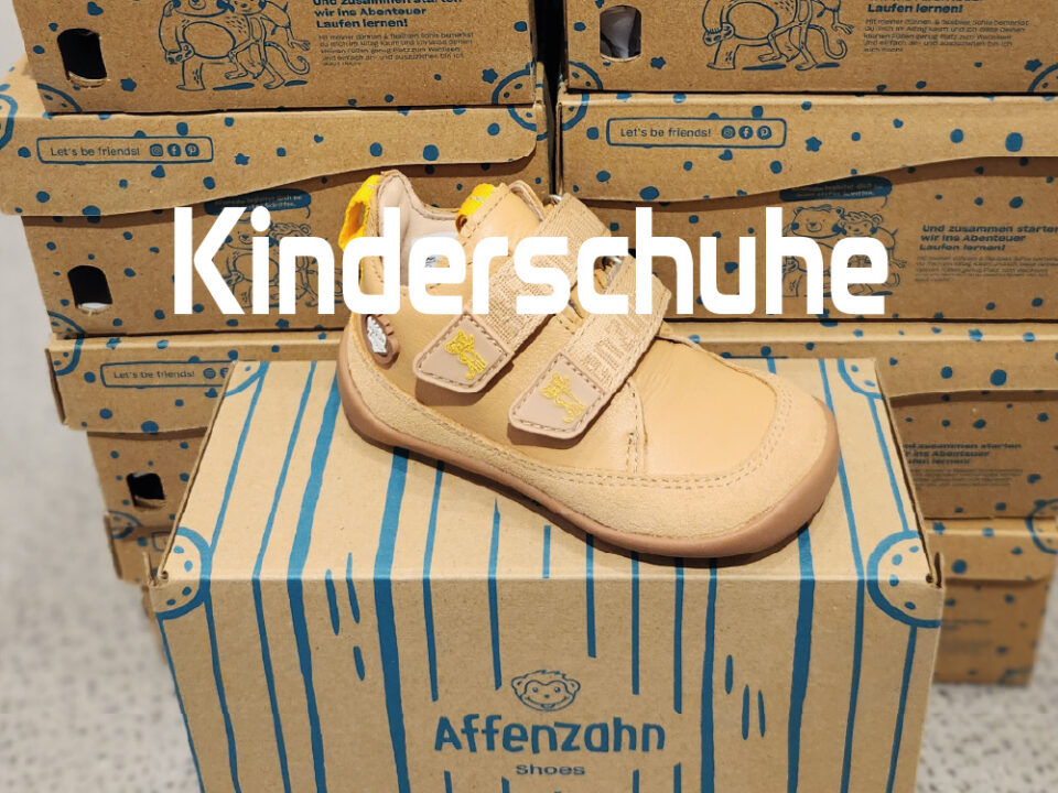 Beiger Affenzahn Kinderschuh mit zwei Klettverschlüssen und brauner Sohle auf einem Schuhkarton, vor gestapelten Kartons mit dem Schriftzug Kinderschuhe.